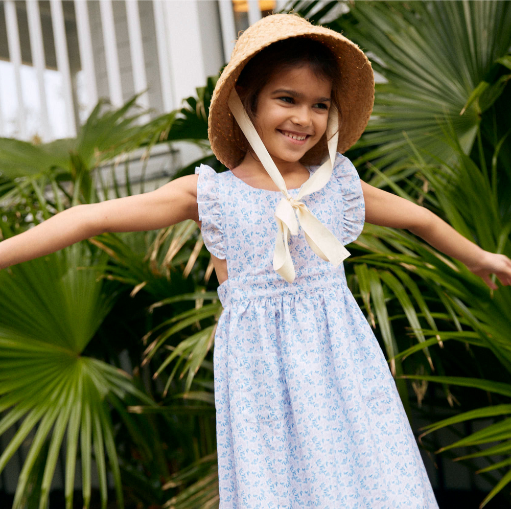 Front view of Briland blue pinafore dress worn by girl, ruffled sleeves, gathered waist, blue floral print, plants background