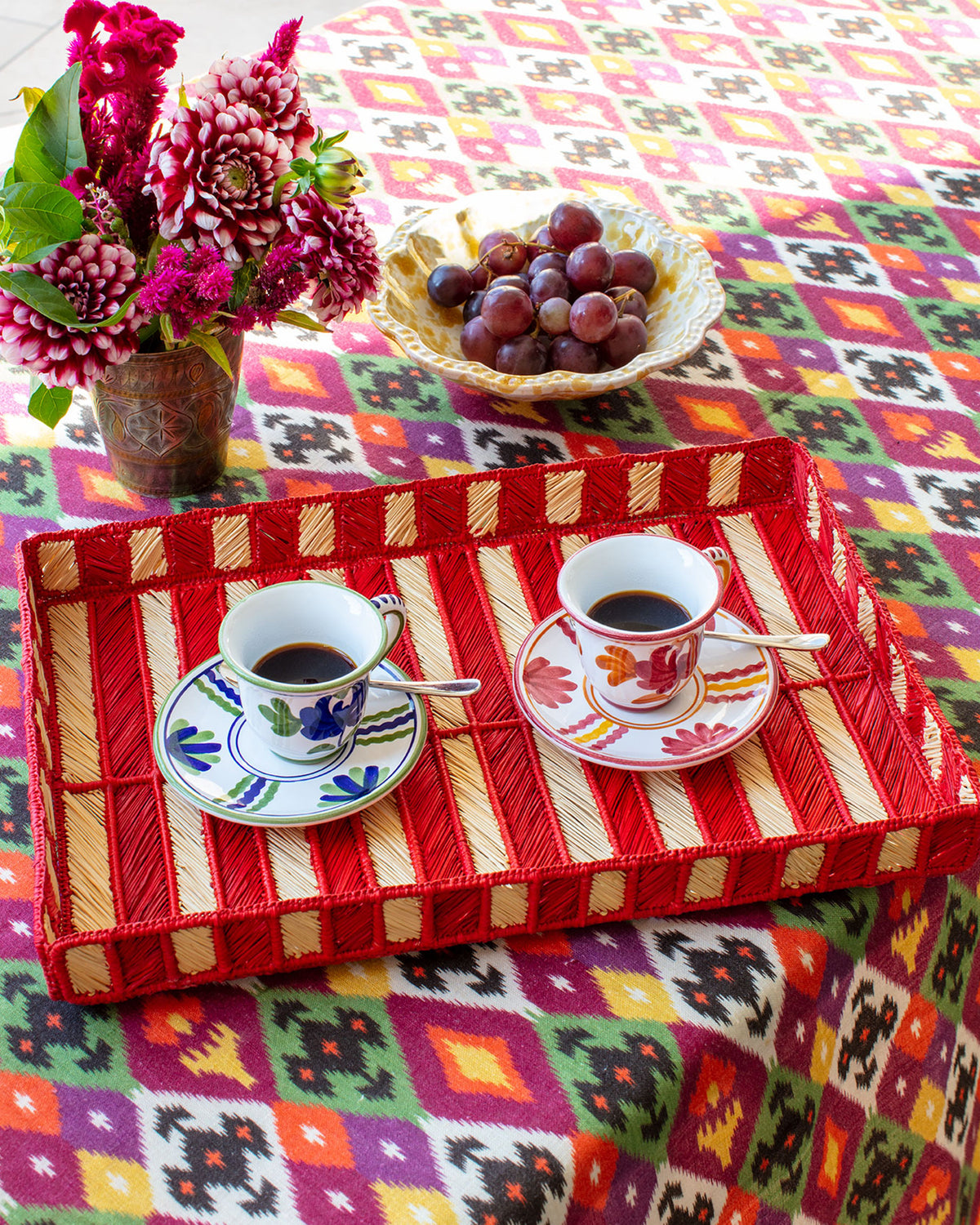 Espresso cups and saucers arranged on a woven tray with flowers, fruit, and colorful patterned textiles