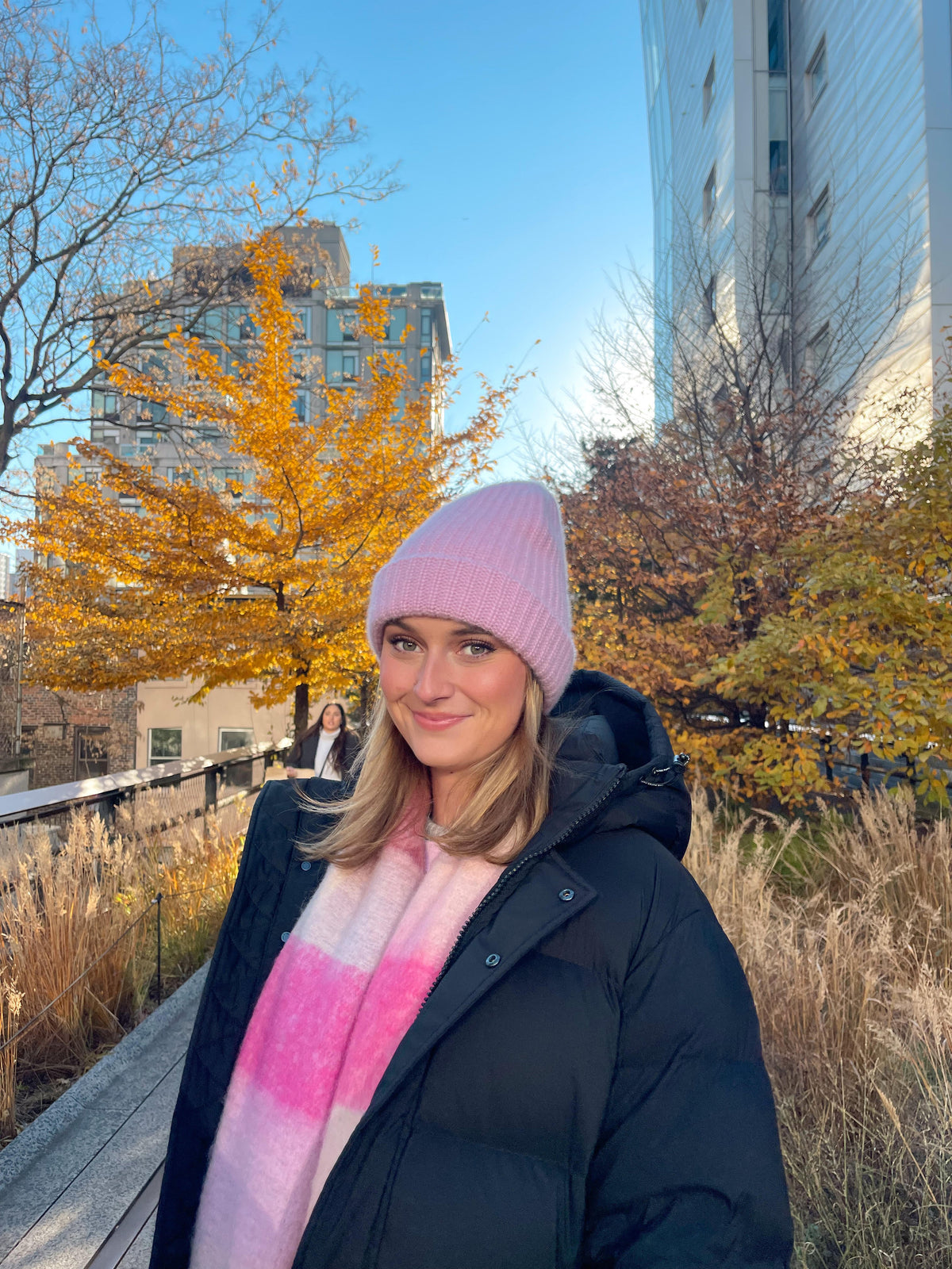 Woman wearing a pink cashmere beanie outdoors among autumn trees and city buildings