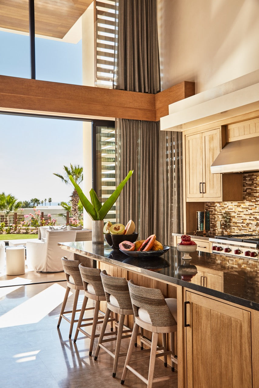 Kitchen island with four woven bar stools and bowls of tropical fruit, open-plan living area with sliding doors opening to an ocean-view patio