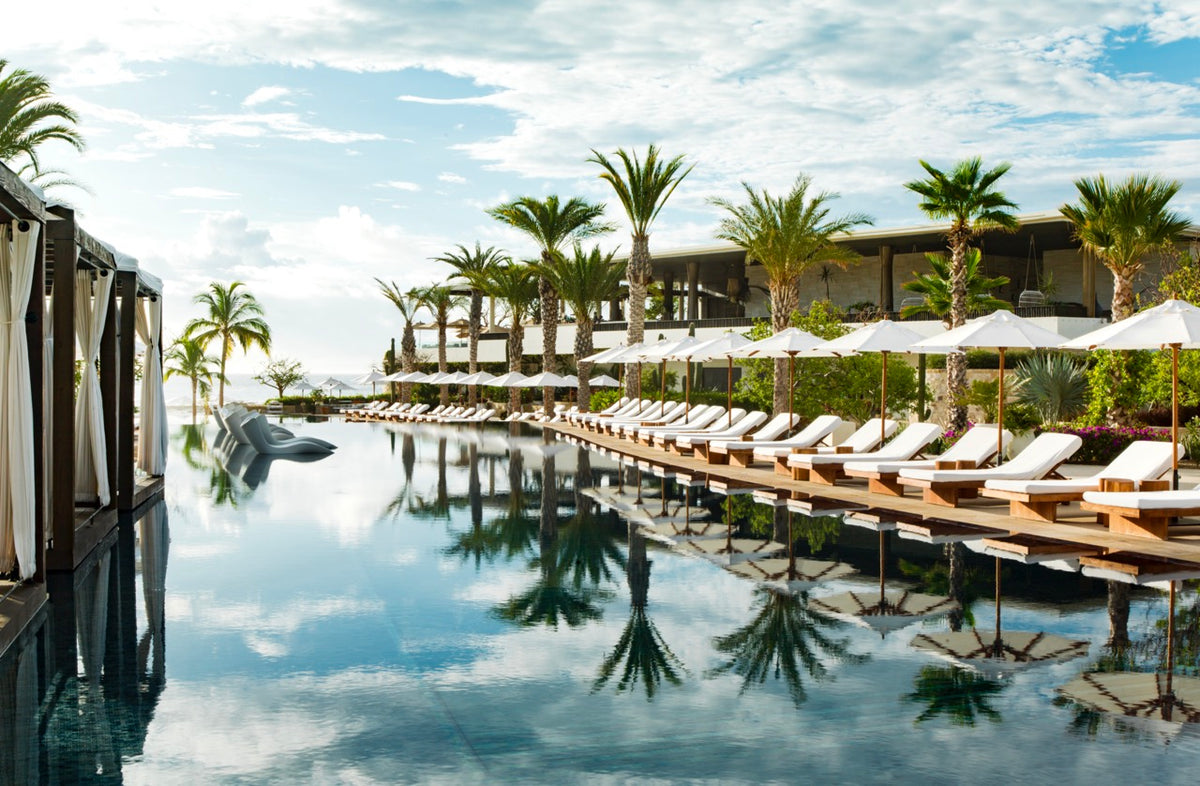 Infinity pool at Chileno Bay lined with white lounge chairs, umbrellas, and cabanas with palm trees reflected in the water and ocean beyond