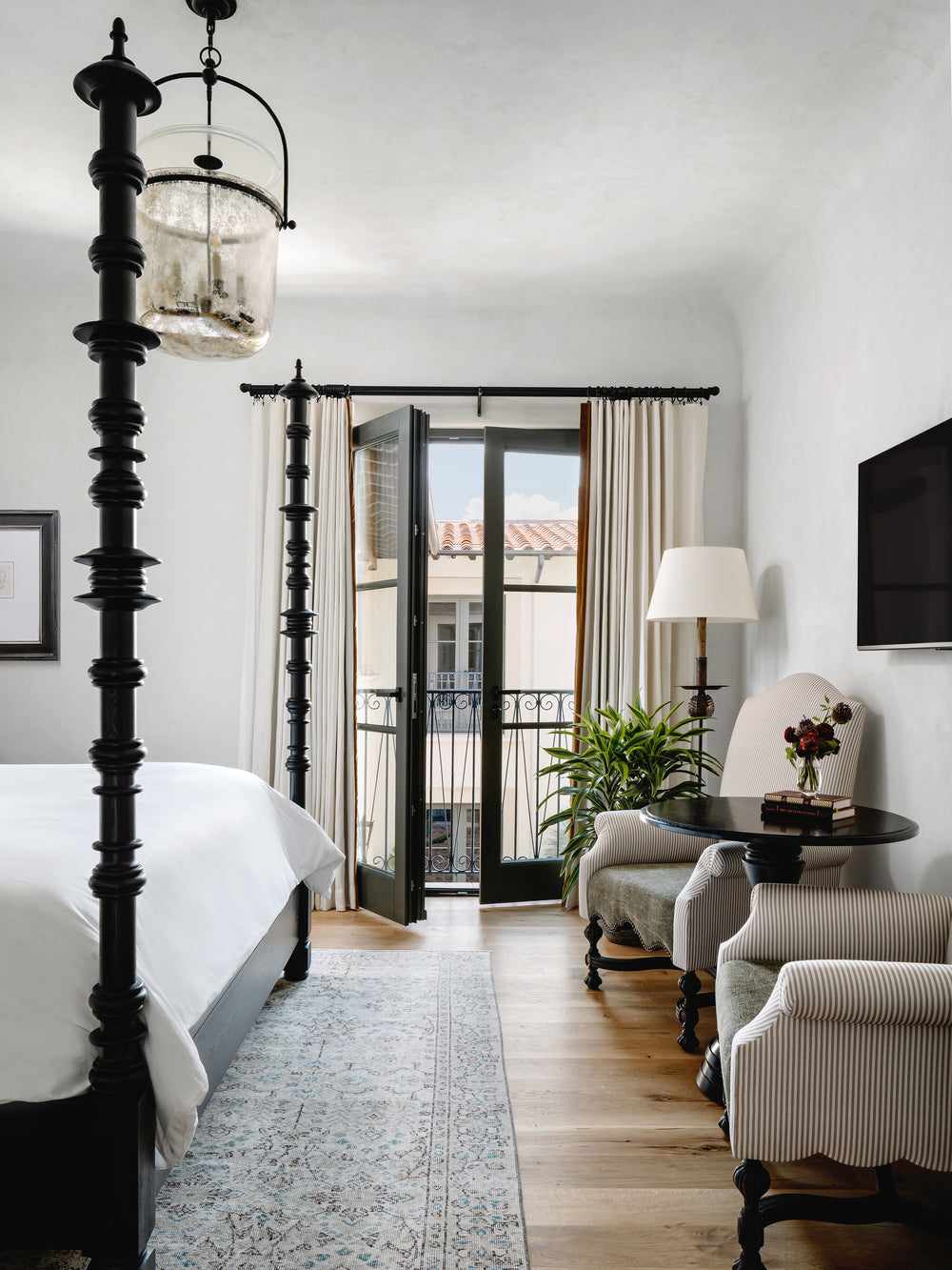 Four-poster hotel bedroom with open French doors to a small balcony, two upholstered armchairs and a round table