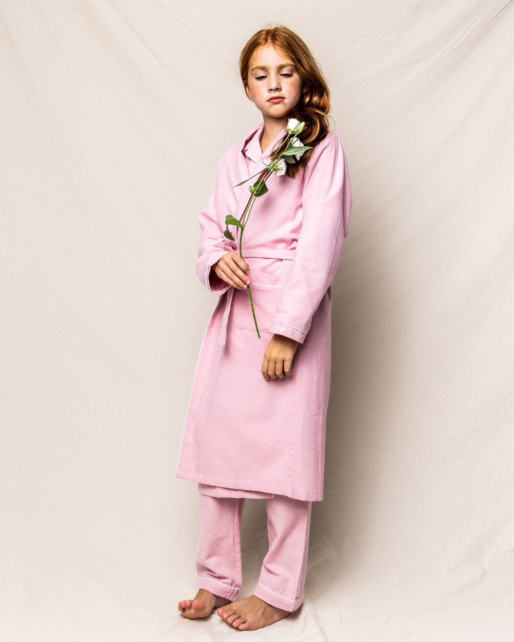 Full-length front view of a child’s pink flannel robe on model showing shawl collar, self-tie belt and side pockets against a neutral backdrop