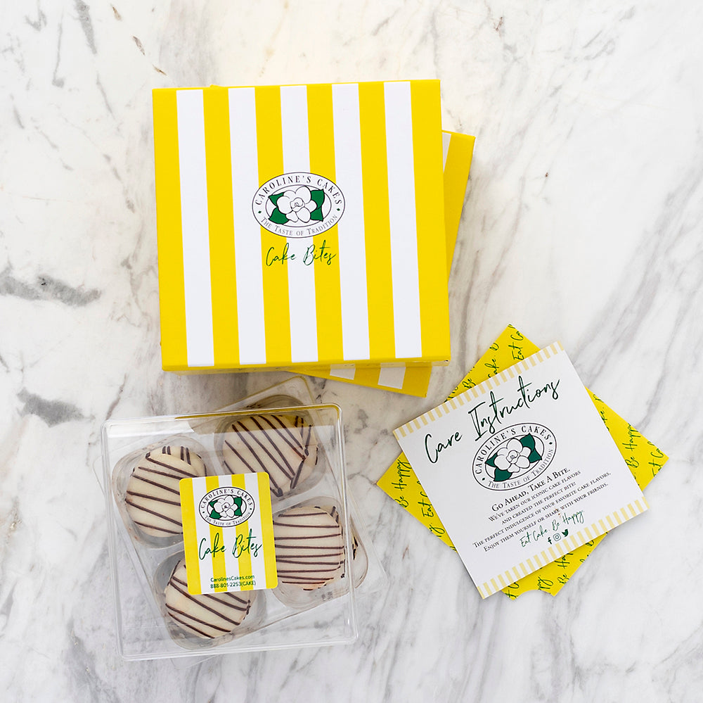 Top down view of four Birthday Cake Bites with chocolate drizzle in a clear box next to a yellow striped gift box on marble
