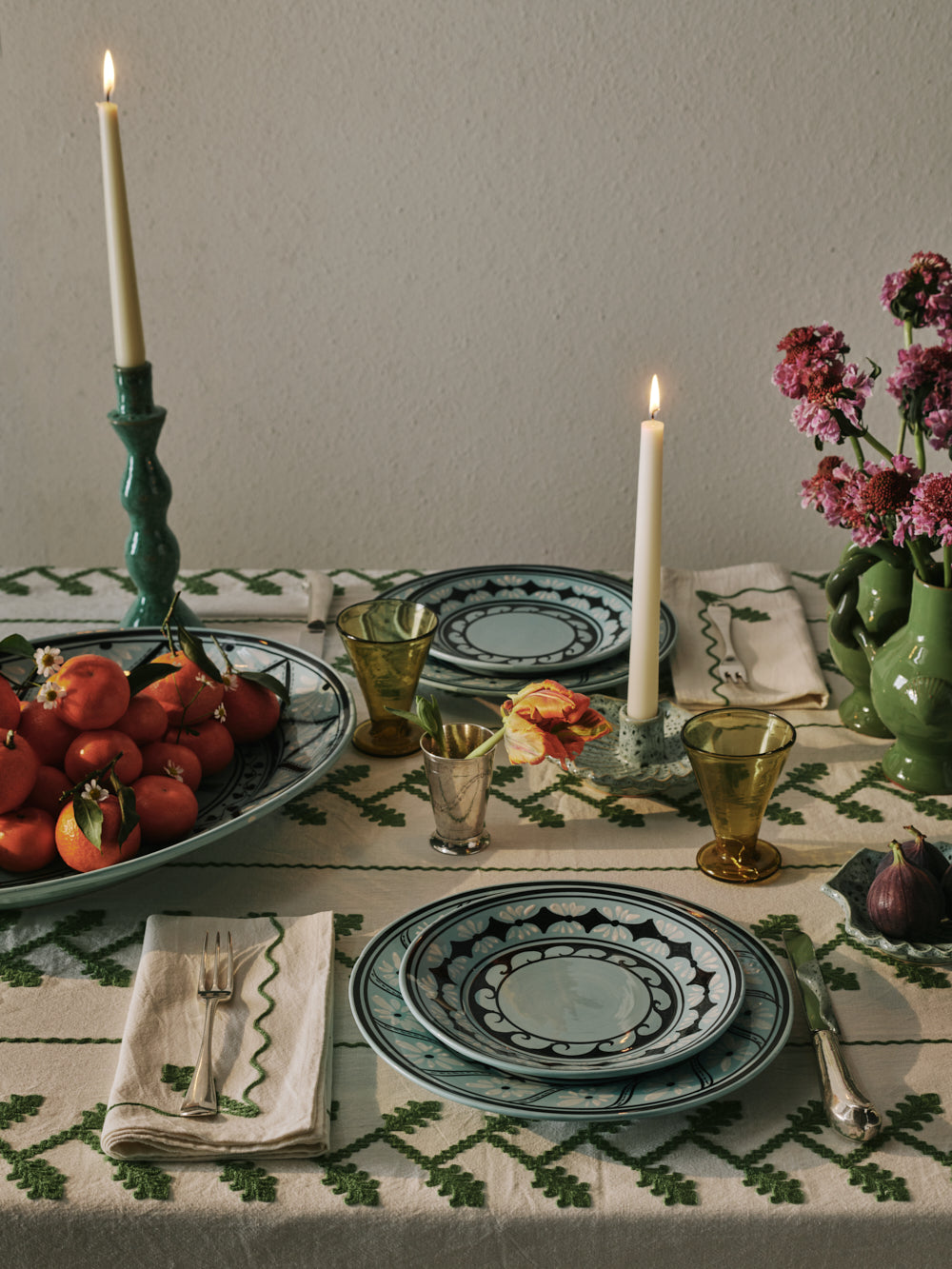 Gigi Aqua dinner plate stacked with matching plate on embroidered tablecloth with lit candles, clementines, and green glassware