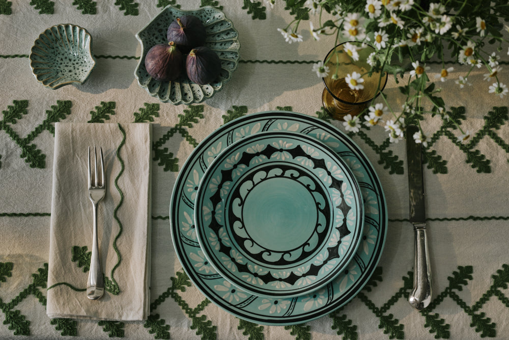 Top-down view of Gigi Aqua dessert plate showing hand-painted aqua glaze and black geometric wave border on embroidered tablecloth