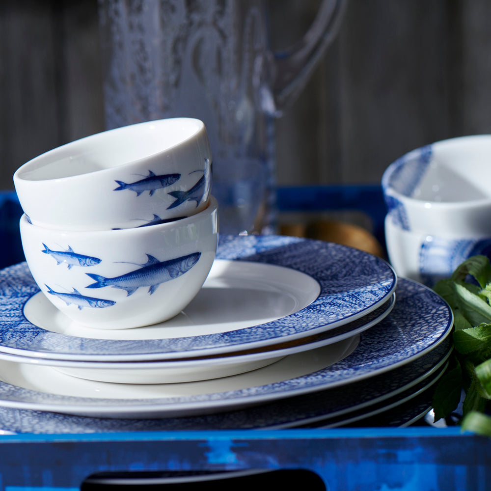 Angled view of School of Fish Snack Bowl showcasing blue fish on creamy porcelain with stacked blue and white dinnerware