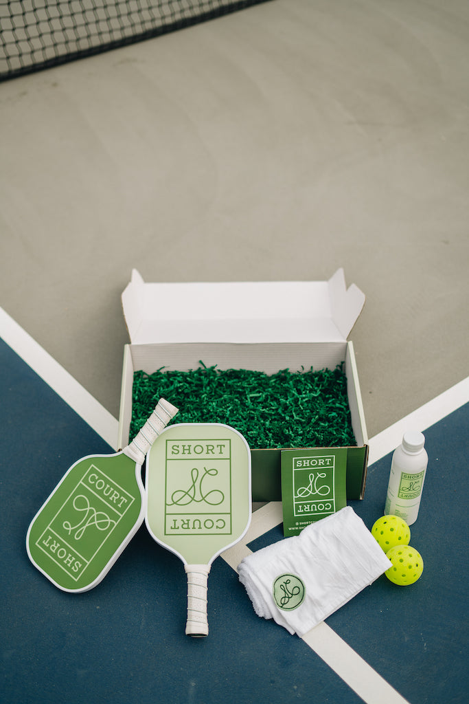 Top-down view of Courtside Collection Doubles box with two Short Court paddles, towel, two pickleballs, and bottle on a blue court