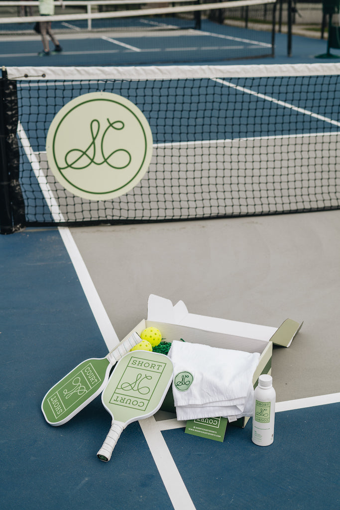 Angled view of Courtside Collection gift box with two Short Court paddles, two balls, folded towel, and a water bottle on a pickleball court