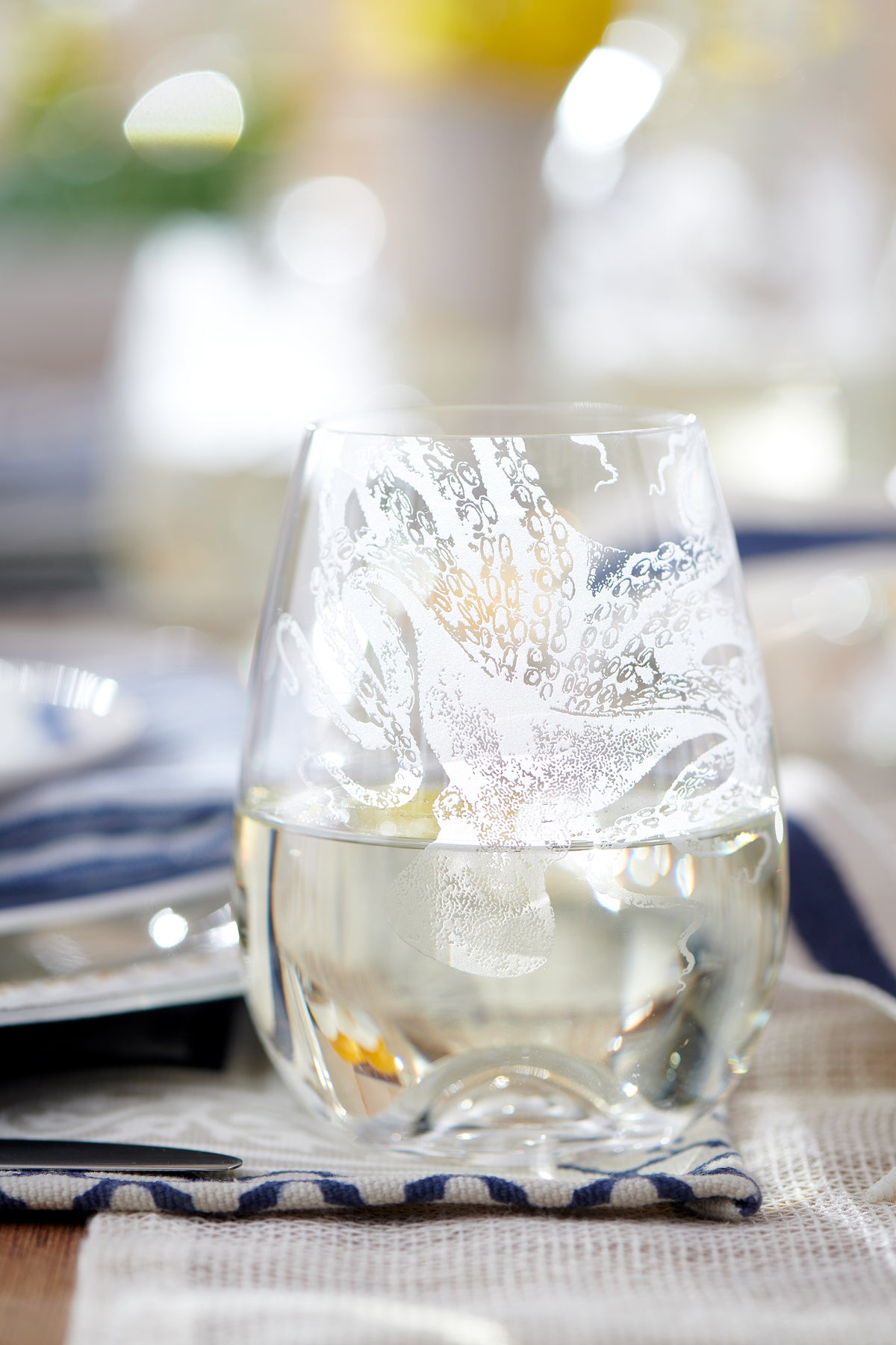 Close-up of Lucy Stemless Wine Glasses, Set of 2 showcasing sand-etched cephalopod design with blurred table setting