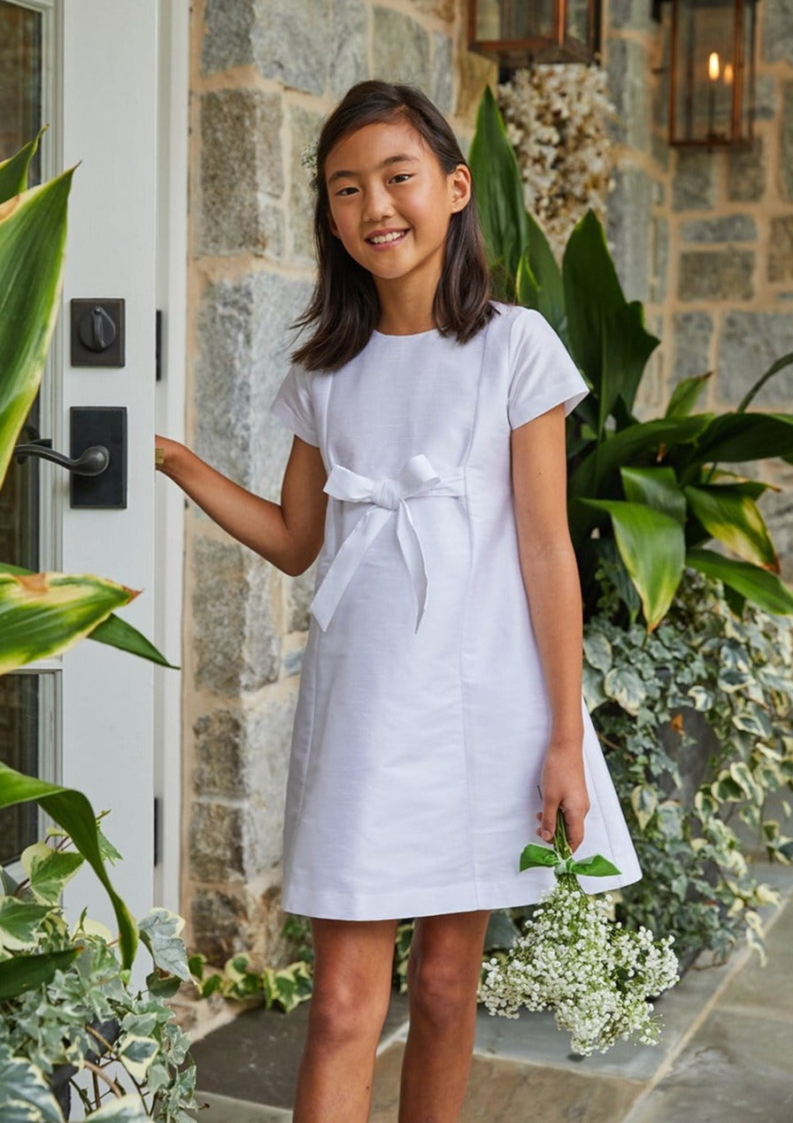 Front view of Cora Dress in Special Occasion White showcasing short sleeves, tied waist bow, and A-line fit on a girl at a stone doorway
