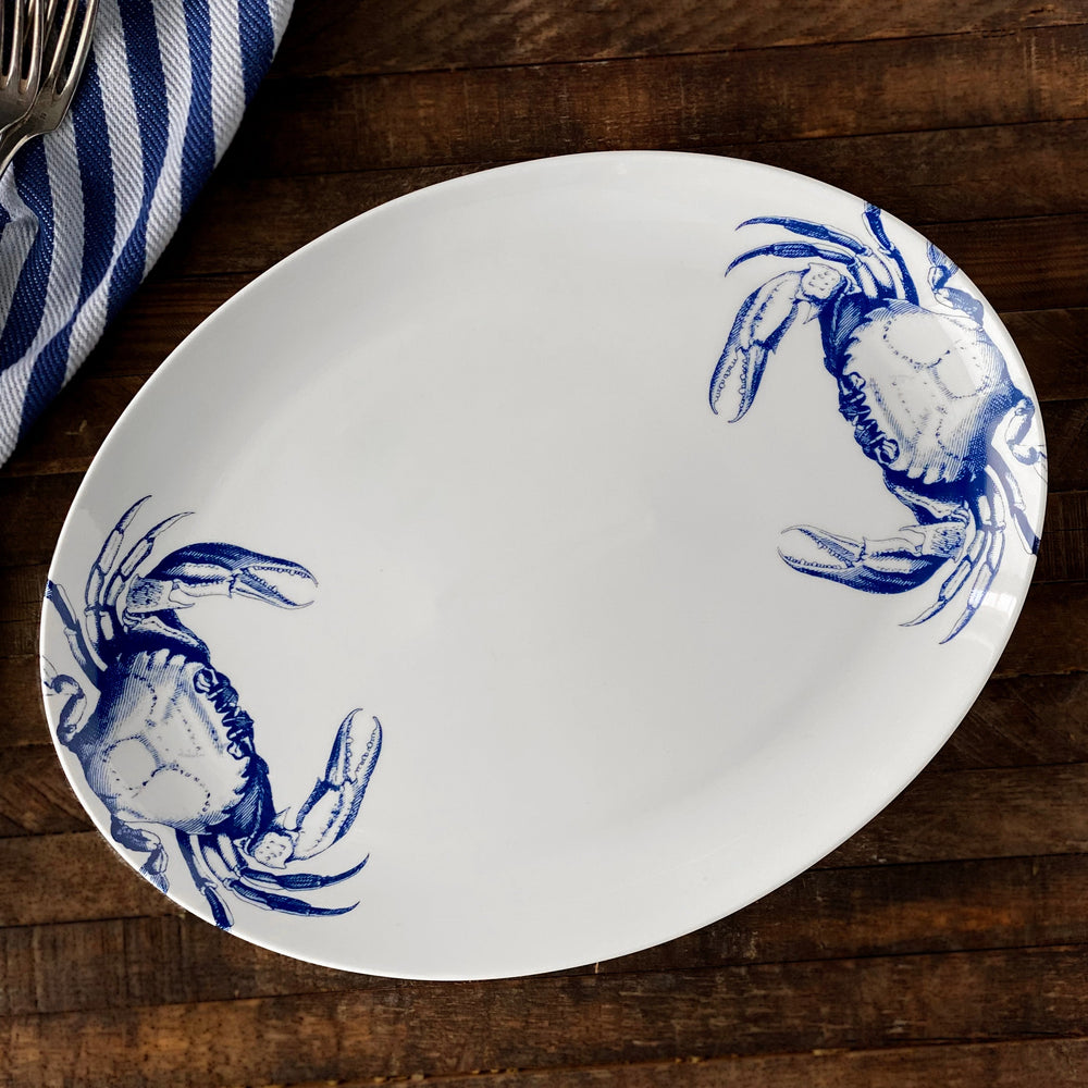 Top-down view of Crabs Blue coupe oval platter showcasing blue crab illustrations on the rim on a wood table with striped napkin