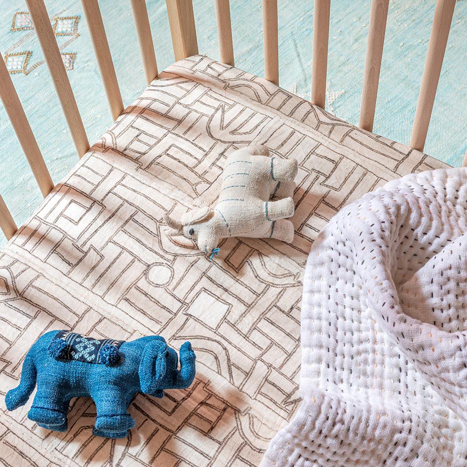 Close-up of Ecru Maze Kuba Cloth Crib Sheet showcasing its geometric pattern on a crib with plush toys and a white blanket