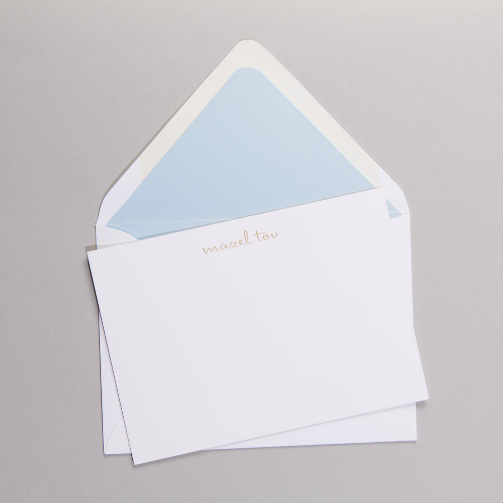 Top-down view of Mazel notecards set showing gold mazel tov lettering and light blue envelope lining on gray background