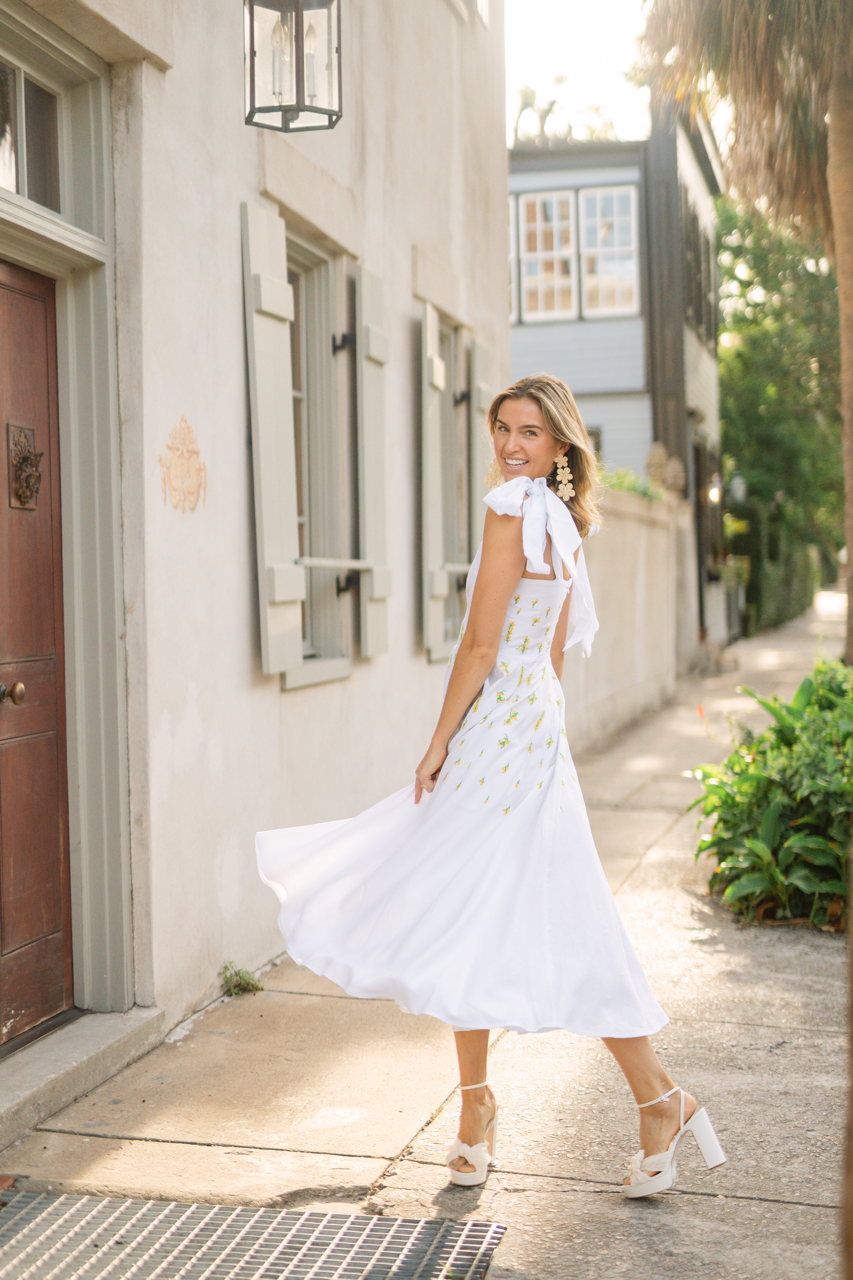Three-quarter view of Mimose Dress on model showing white linen, tie shoulder bows and small floral embroidery, sidewalk setting