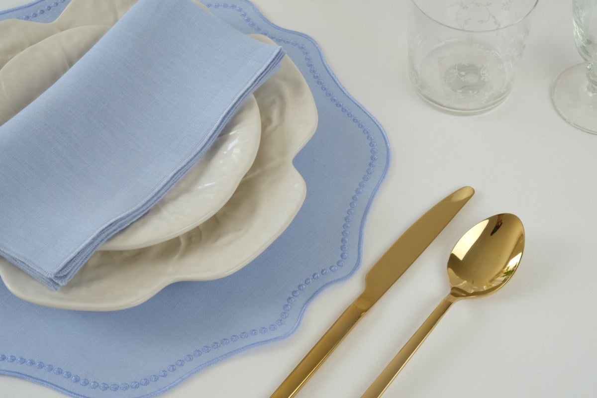Top-down view of blue linen placemat with dotted embroidered border and scalloped edge under stacked cream plates, a light-blue napkin, and gold flatware