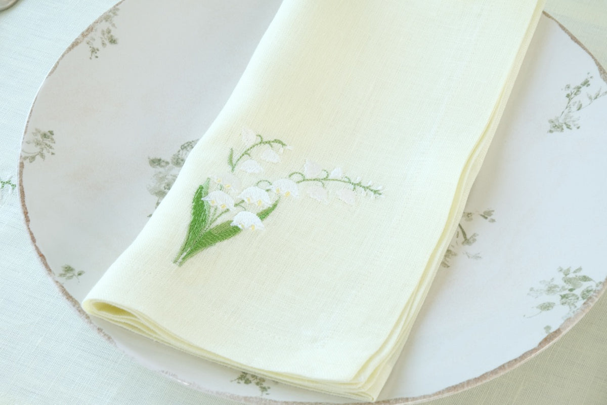 Top-down view of a folded light-green linen napkin with embroidered lily-of-the-valley sprig on a floral dinner plate