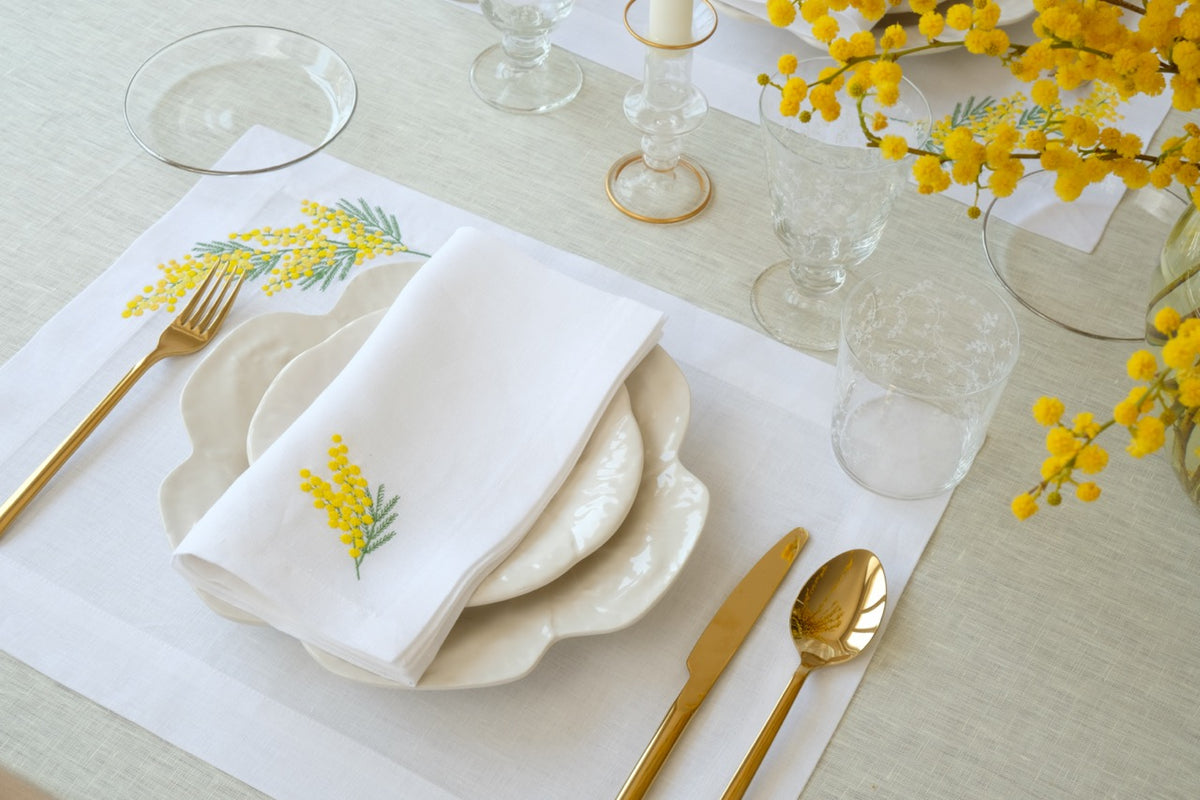 Top-down view of Mimosa napkin and placemat set showcasing yellow mimosa embroidery on white linen with gold flatware