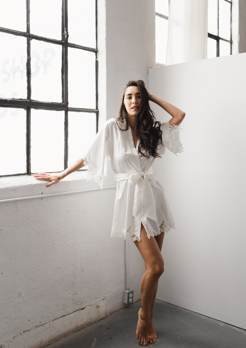 Three-quarter view of Cara silk cotton short bridal robe on a model showcasing scalloped lace-trimmed sleeves and hem with a tied sash in a window-lit studio