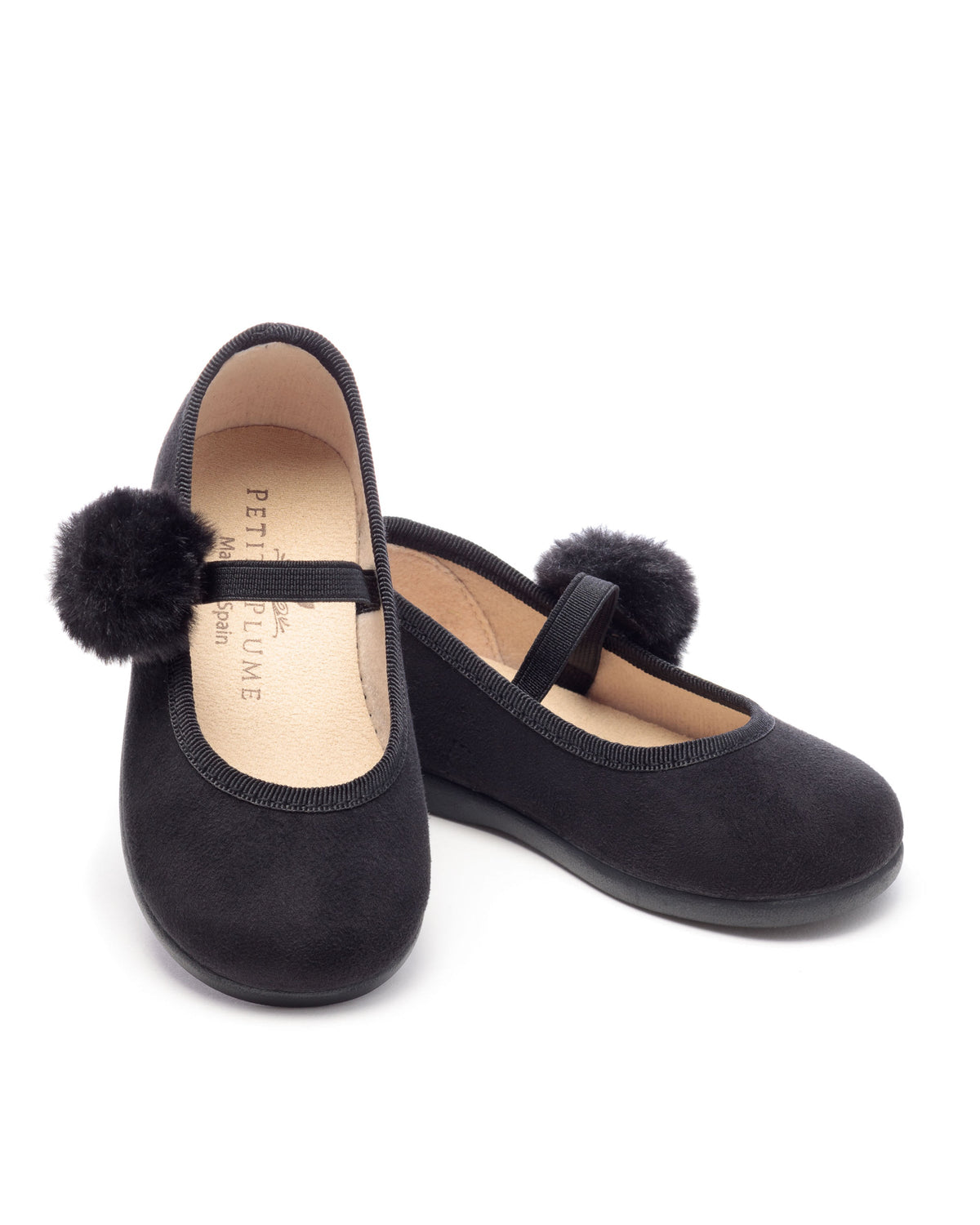 Three-quarter view of The Delphine Slipper in Black Suede with twin pom poms and Mary Jane straps on white