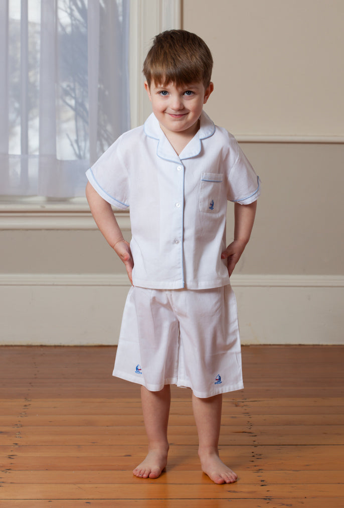 Front view of boys white short set on model with Bermuda shorts and blue sailboat embroidery on wood floor