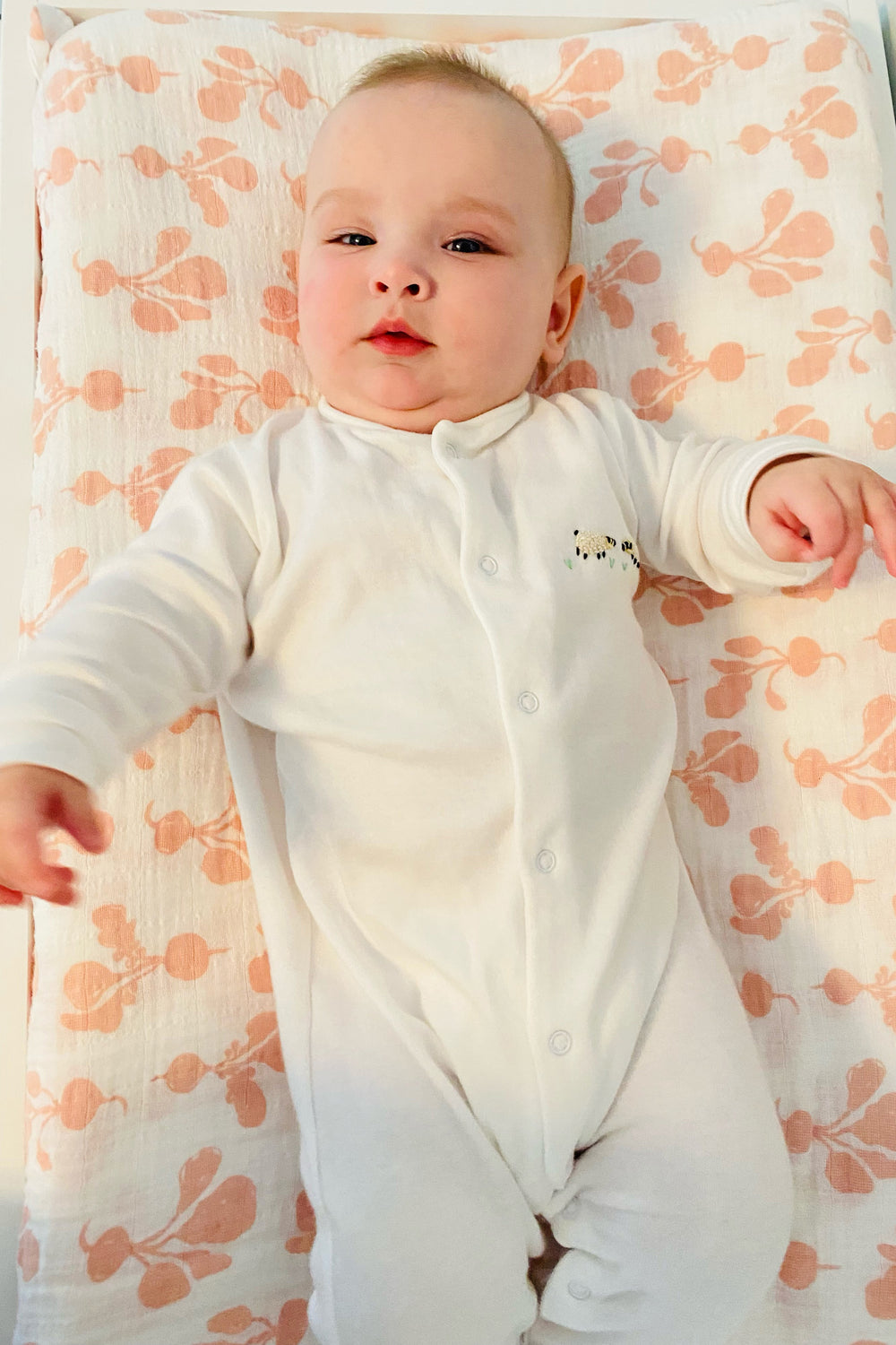 Front view of baby footie worn by an infant showcasing cream cotton and small embroidered sheep on a pink floral blanket