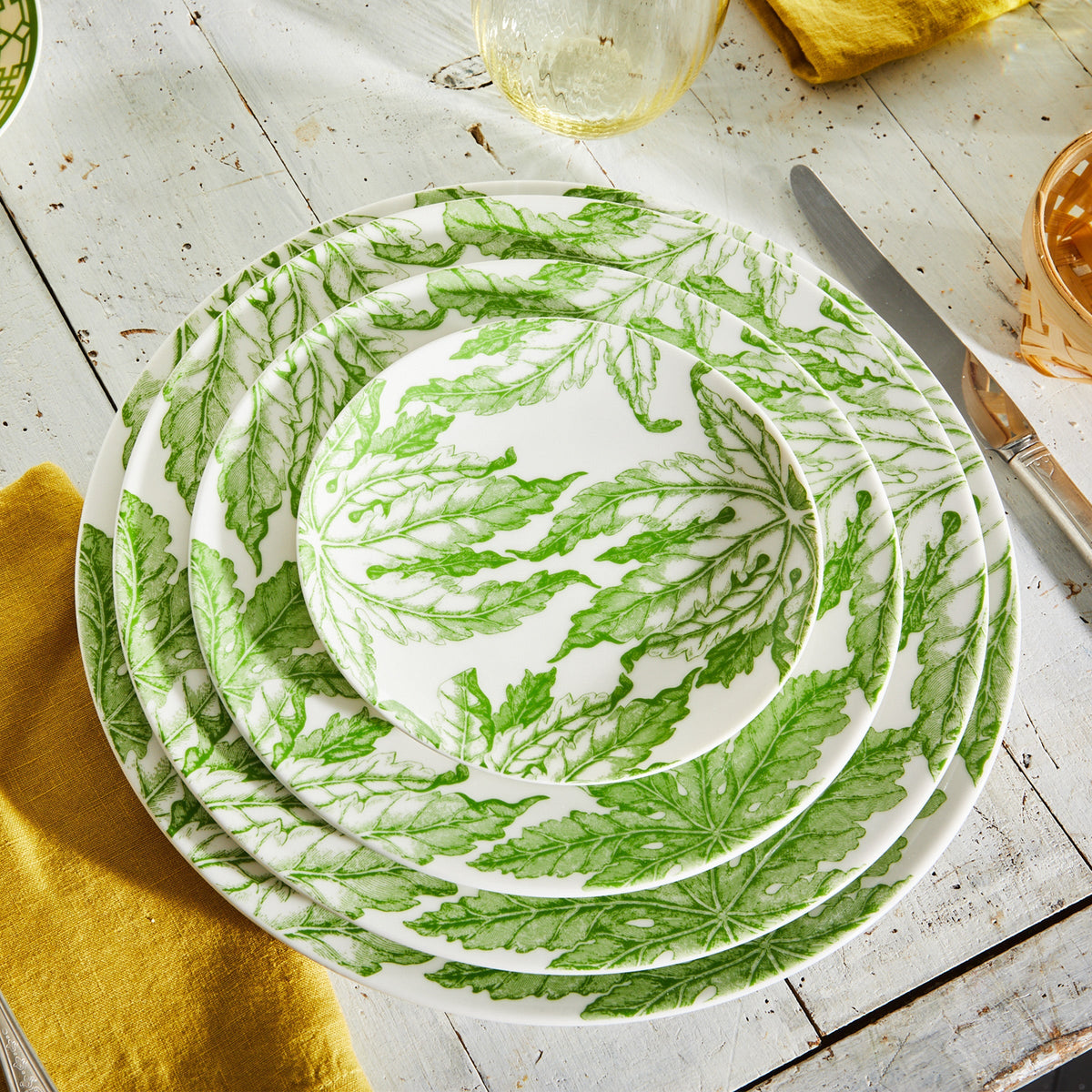 Top view of Freya Rimmed Charger showcasing green floral pattern on creamy white porcelain with stacked dinnerware on a rustic table