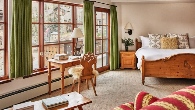 Cozy alpine-style hotel room with wooden bed, bedside table and desk by large windows with green curtains opening onto a small balcony view