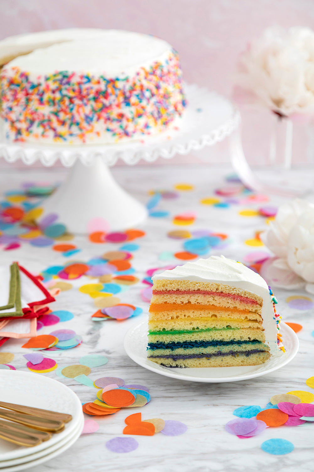 Front view of Sprinkle Rainbow Cake slice showcasing seven rainbow-colored layers and white buttercream frosting with rainbow sprinkles on a marble table