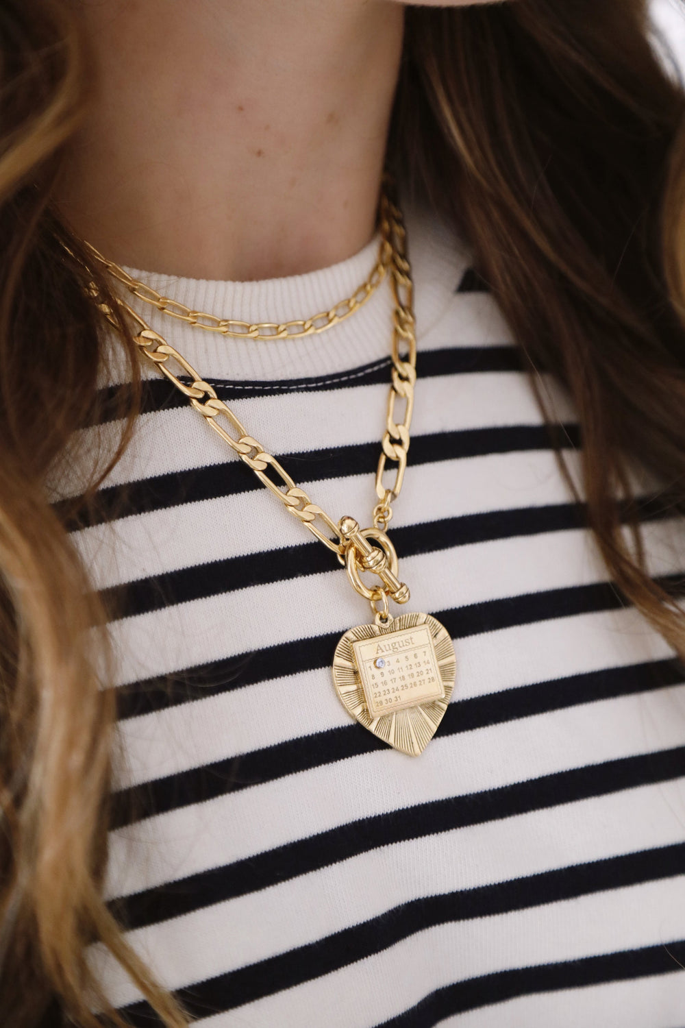Close-up of The Best Day necklace showcasing layered gold Figaro chains and a heart pendant engraved with an August calendar on a striped top