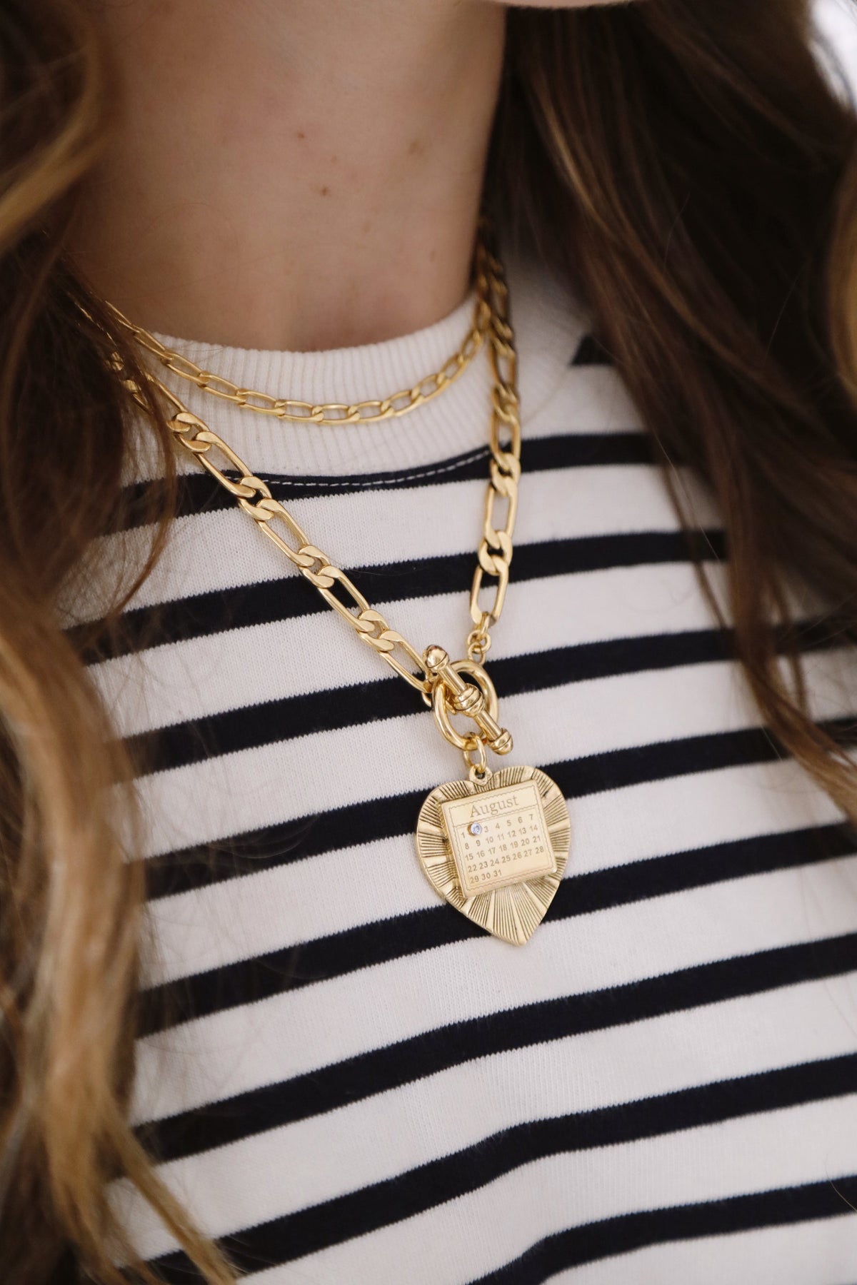 Close-up of The Best Day necklace showcasing layered gold Figaro chains and a heart pendant engraved with an August calendar on a striped top