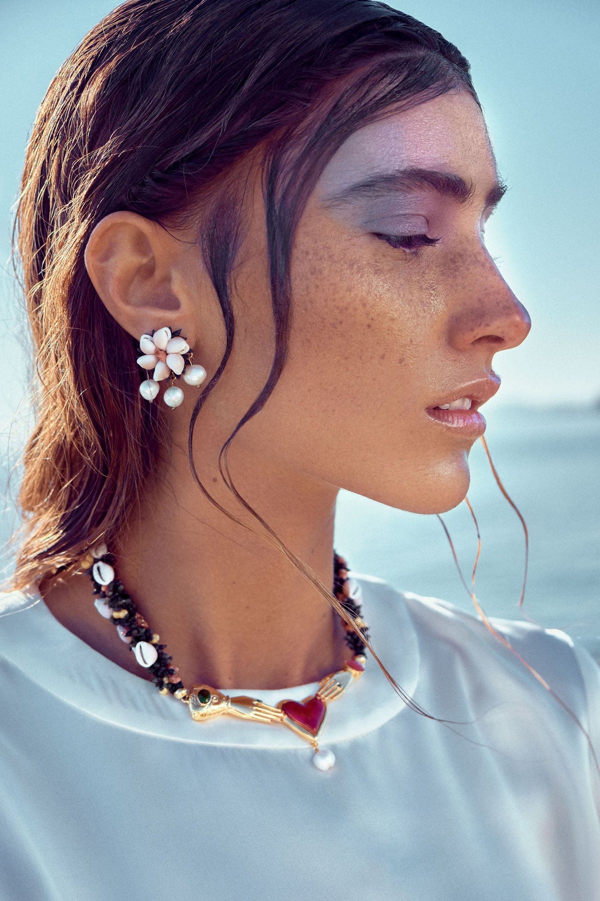 Side view of Shella Earrings showcasing white porcelain petals and natural Baroque pearls against a blue outdoor background