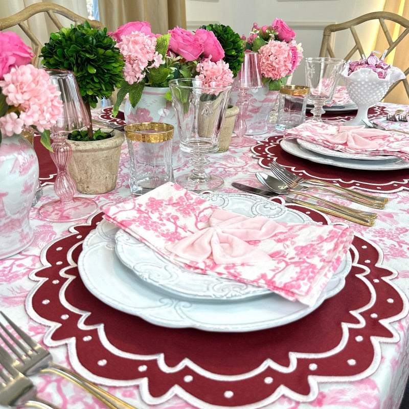 Pink Toile de Jouy napkins folded on white plates in a styled table setting with pink flowers and gold-rim glasses