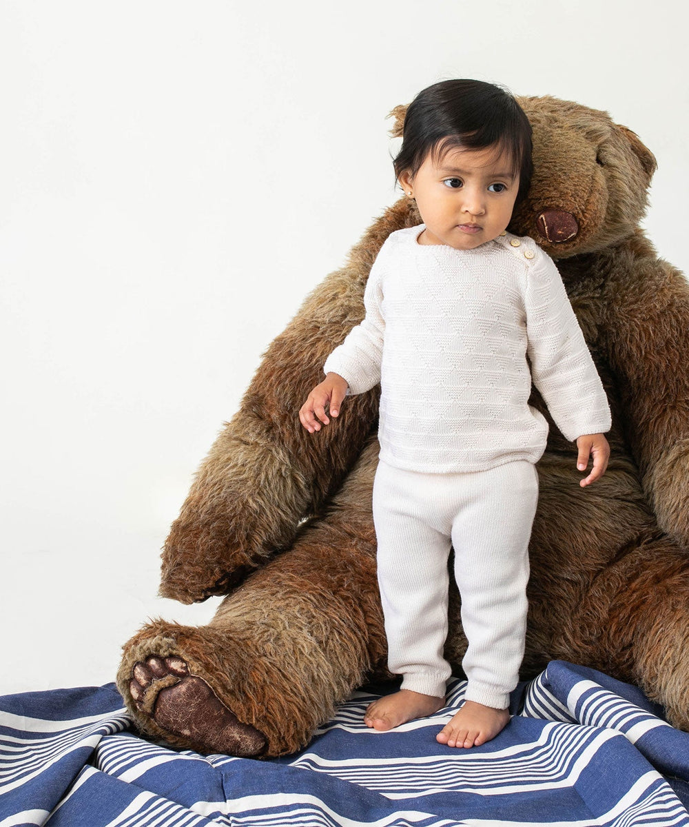 Front view of Baker baby pant in blue on toddler model showing knit texture and ribbed cuffed ankles with large teddy bear backdrop