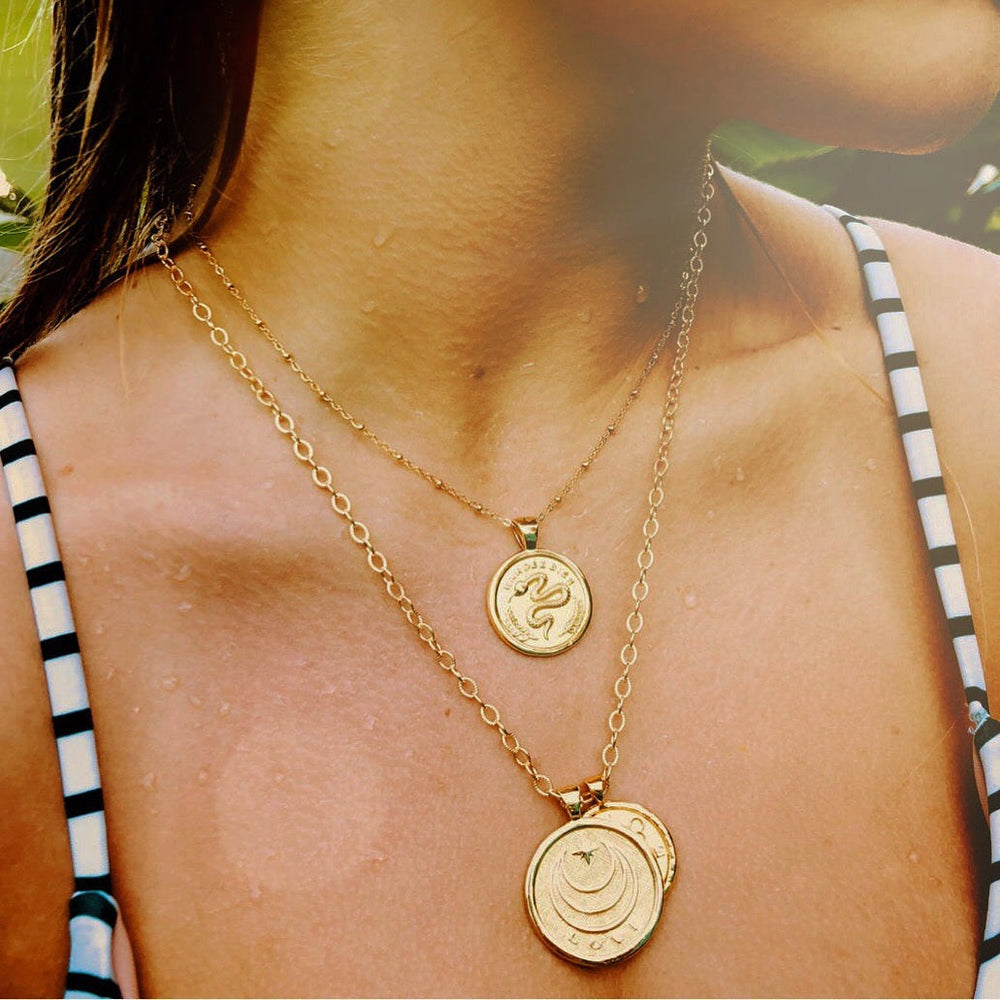 Close-up of Protect JW small gold coin pendant on thin chain layered with larger gold coin necklace on model’s collarbone