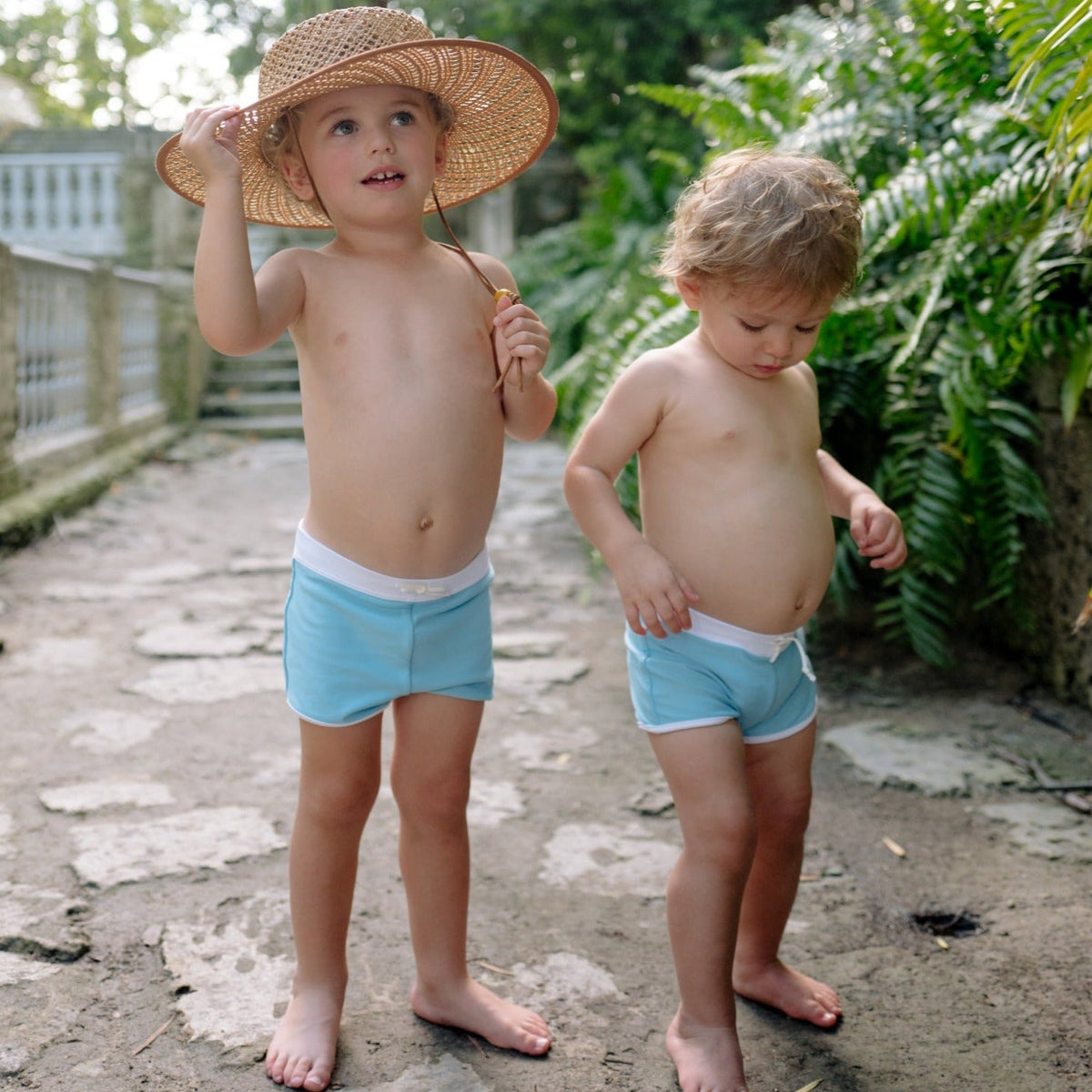 Two young children wearing light blue Boys Lagoon briefs walking on a sunlit stone path near greenery, one holding a straw hat