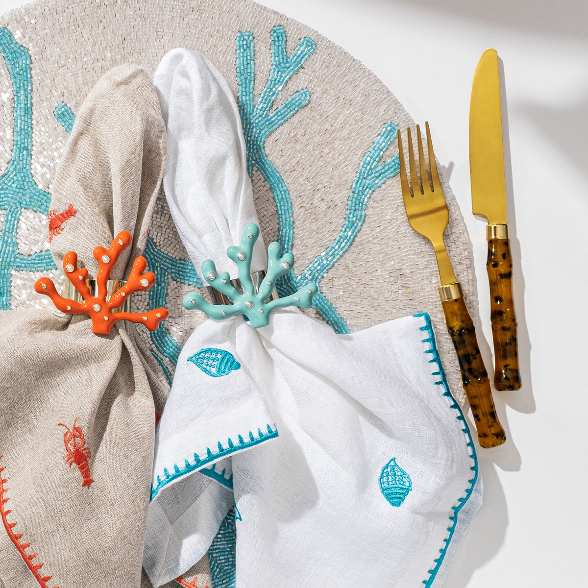Top view of coral-shaped napkin rings (turquoise and orange) on embroidered linen napkins with gold flatware