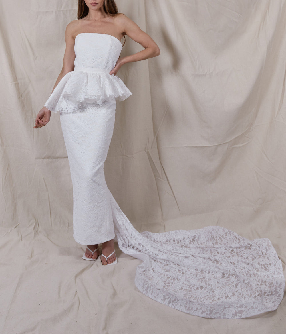 Front view of Maeve Lace Gown showing strapless corset, peplum, fitted ankle-length silhouette and lace train on beige backdrop