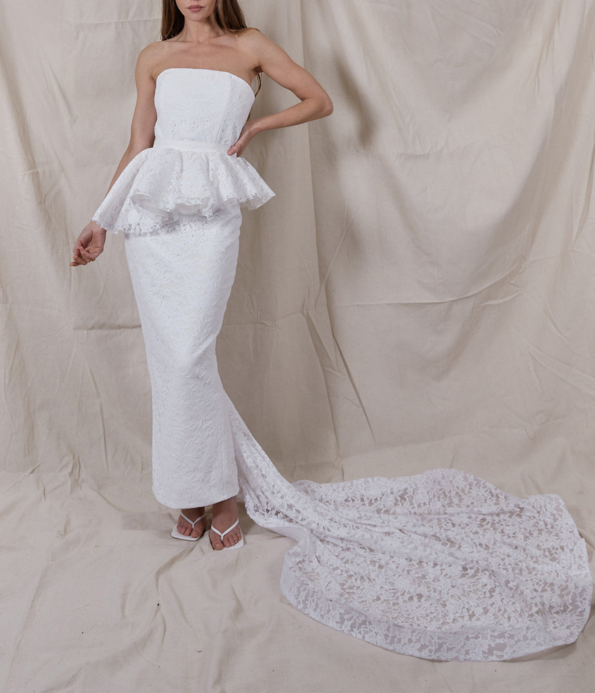 Front view of Maeve Lace Gown showing strapless corset, peplum, fitted ankle-length silhouette and lace train on beige backdrop