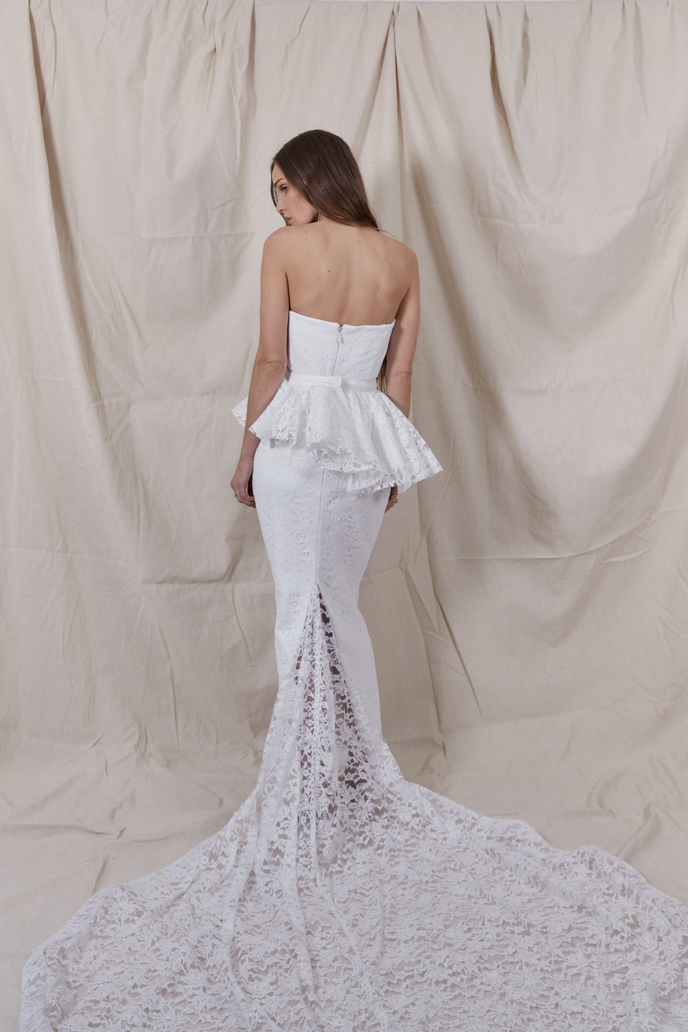 Back view of Maeve Lace Gown showcasing strapless corset, detachable peplum, and lace train against a beige fabric backdrop