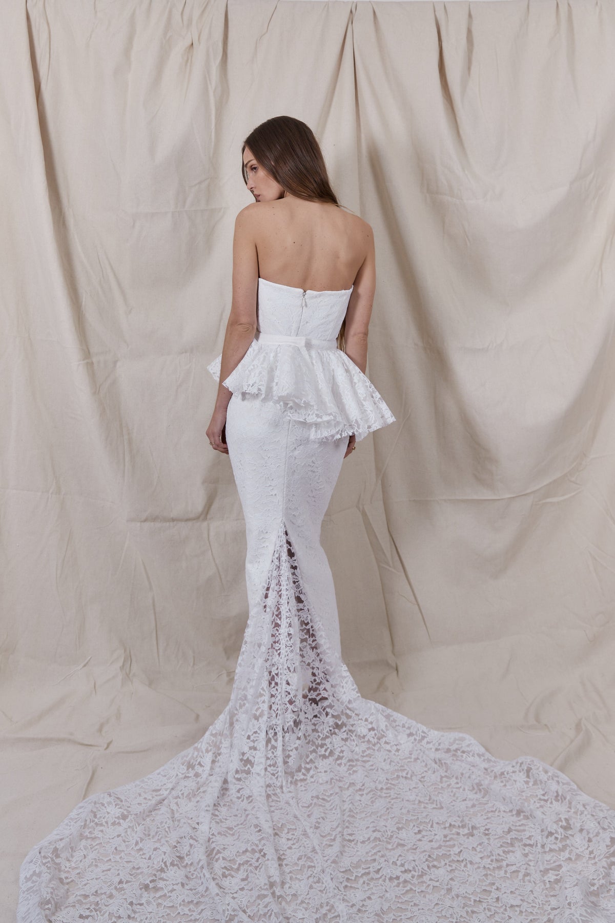 Back view of Maeve Lace Gown showcasing strapless corset, detachable peplum, and lace train against a beige fabric backdrop