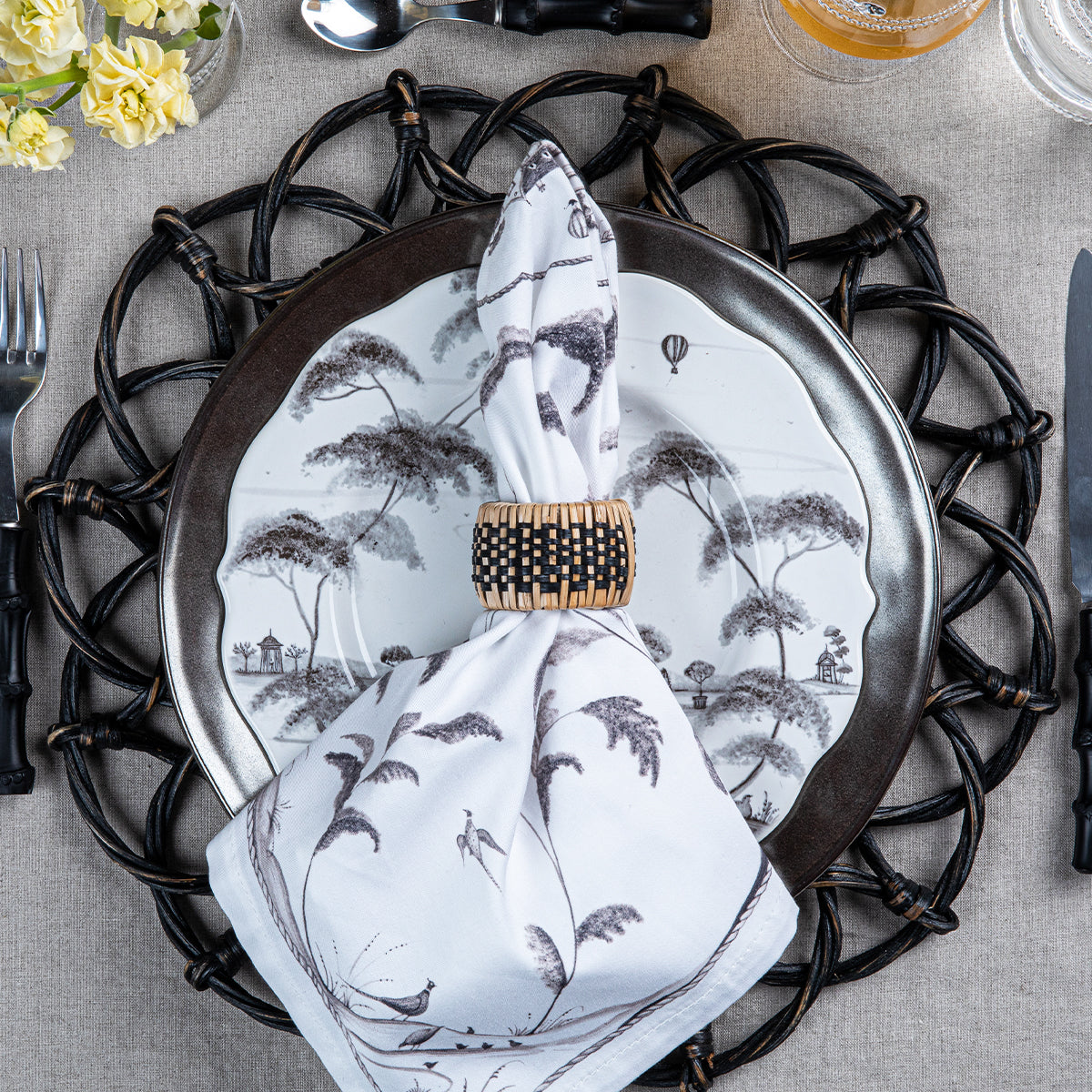 Top view of Woven Napkin Ring showcasing hand-woven rattan and black detailing with a patterned napkin on a table setting