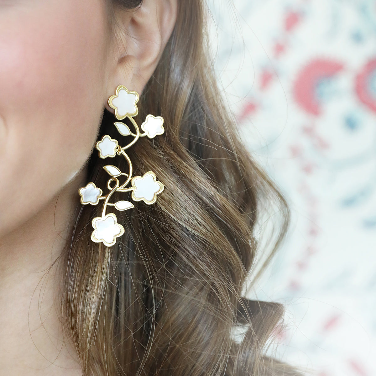 Close-up of ear wearing Lilia Chandelier earring showcasing white mother-of-pearl flower motifs and gold vermeil vine on a light patterned background