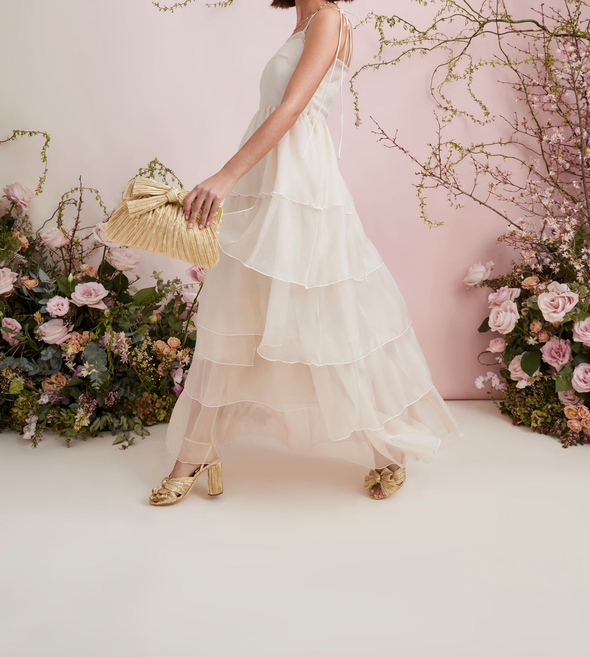 Full length view of Camellia Bow Heel in Gold showcasing gold heels with a layered white dress and floral backdrop