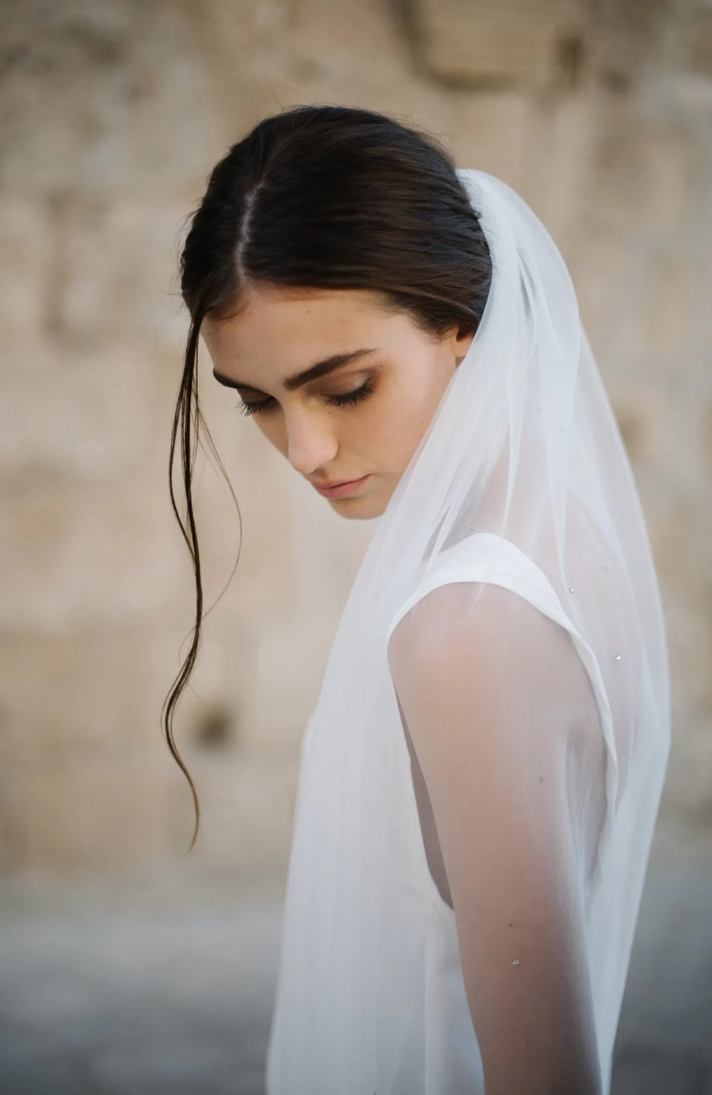 Side view of model wearing Lucca crystal wedding veil, sheer silk tulle with scattered Swarovski crystals, stone background
