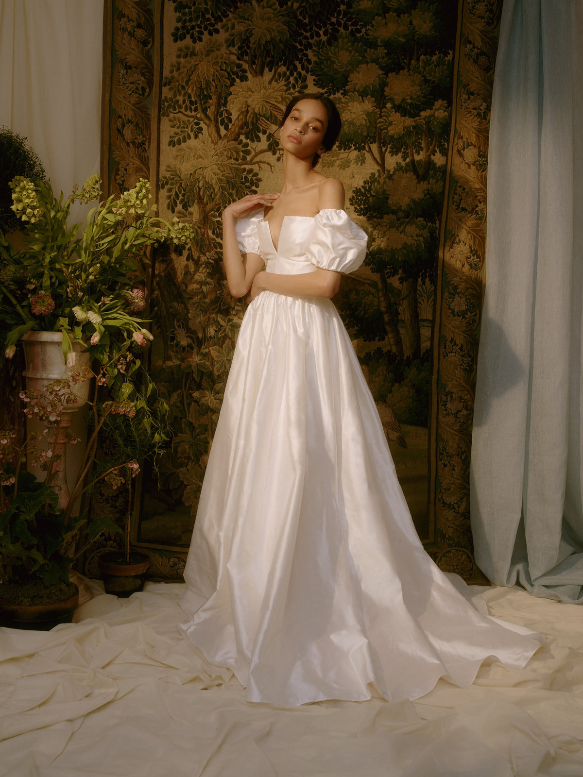 Front view of Yvetta Duchesse Satin Gown showcasing off-the-shoulder puff sleeves and full skirt with floral tapestry background
