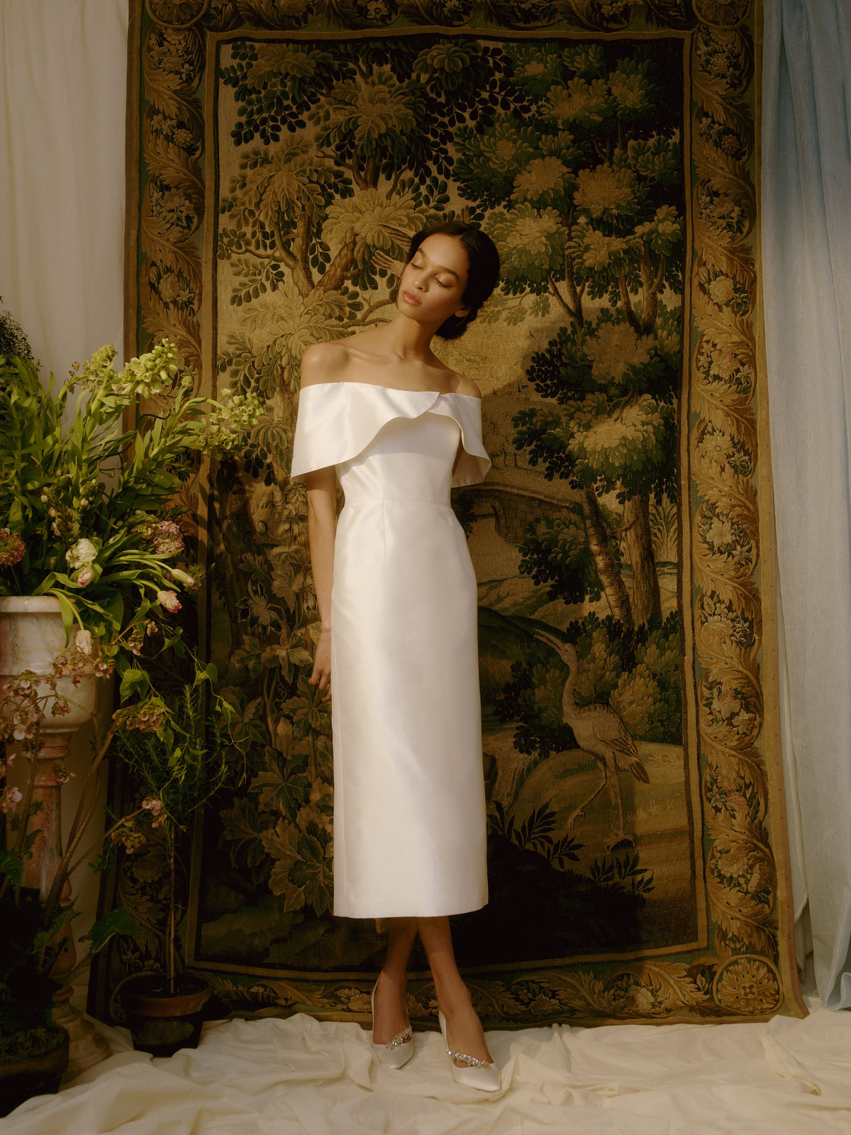 Front view of the slim-fit Eveline White Silk Dress showcasing off-the-shoulder sleeves and scalloped neckline with floral tapestry background