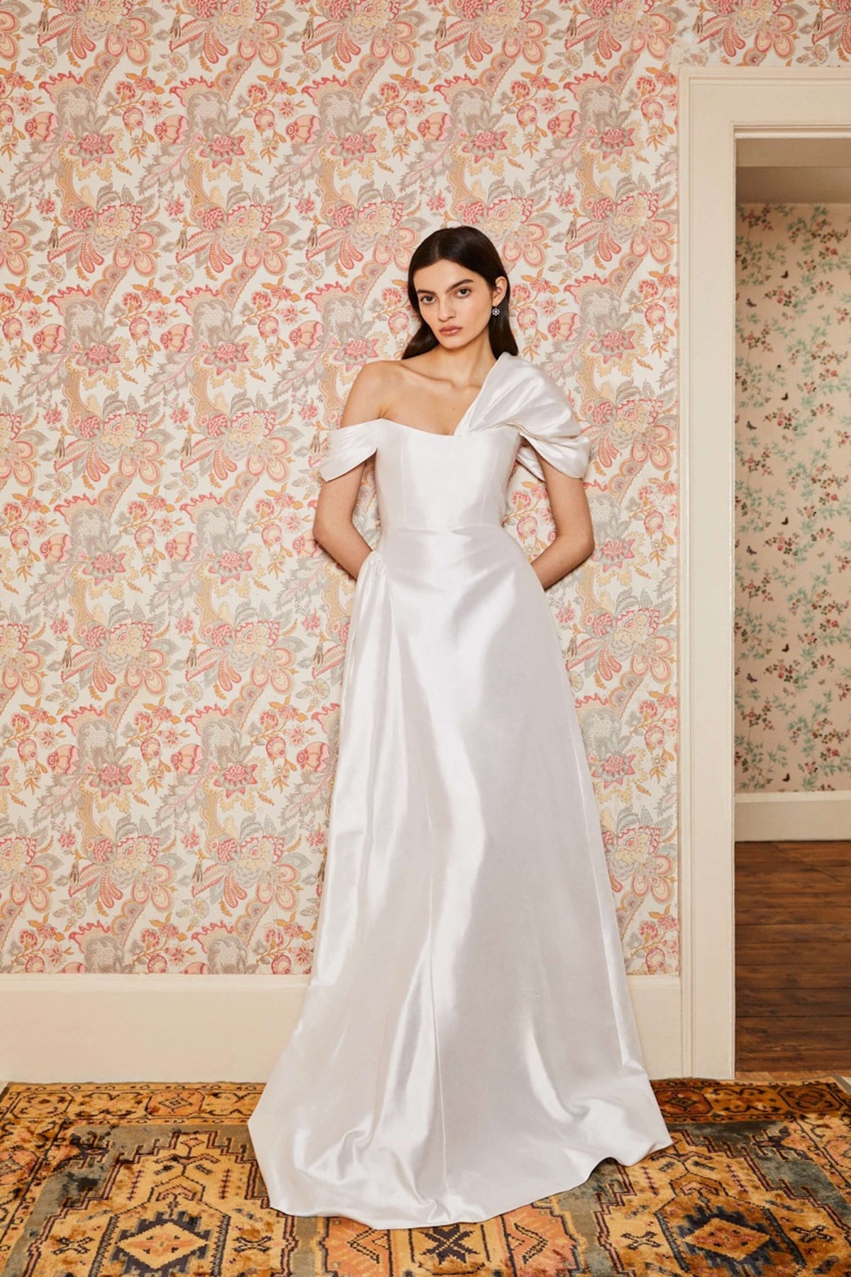 Front view of Edith White Silk Asymmetric Drape Shoulder Gown showcasing gathered waist and scooped neckline with floral wall