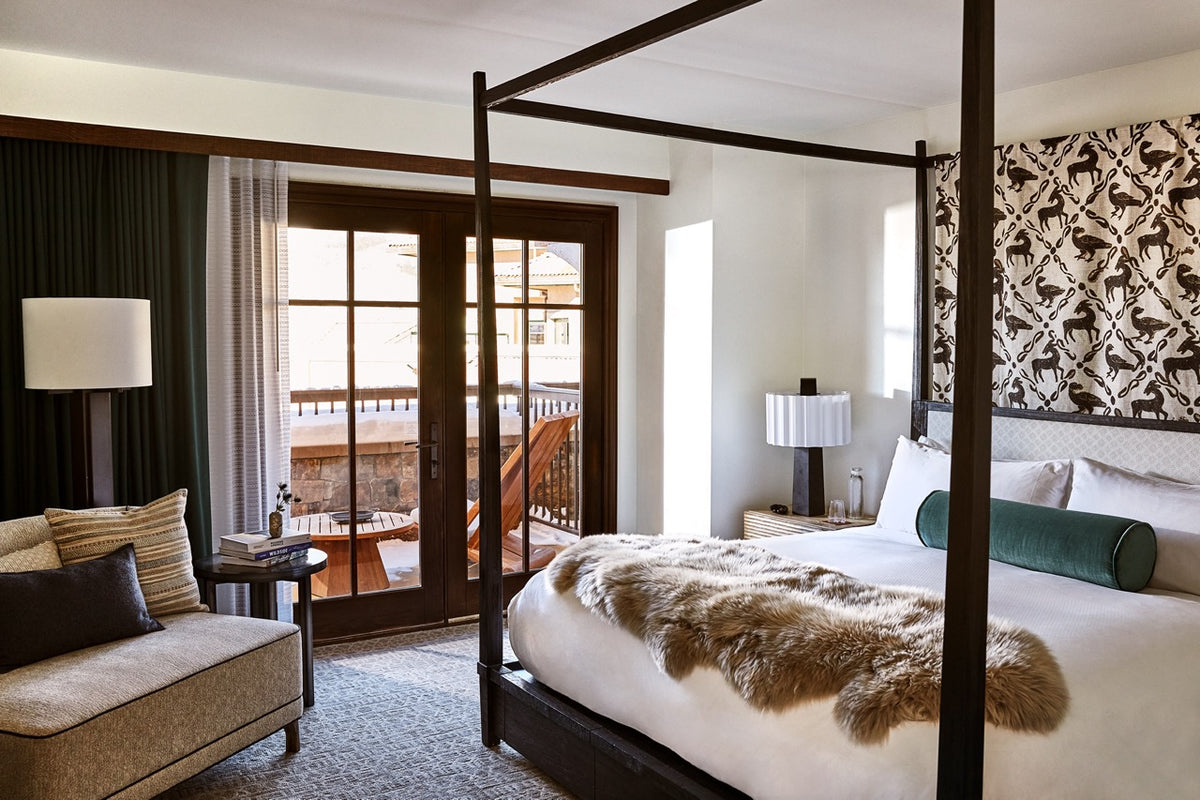 Four-poster king hotel room with fur throw on bed, lounge seating, bedside lamps, and balcony with wooden Adirondack chairs