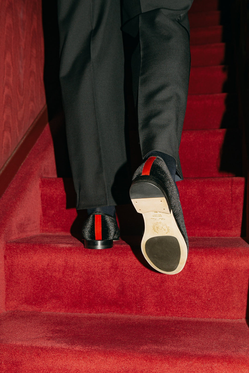 Rear view of Men’s Black Velvet Slipper II showcasing red heel stripe and velvet shimmer on red carpeted stairs